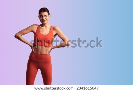 Young sporty female wearing red tight sportswear standing with hands on waist against gradient background and looking away
