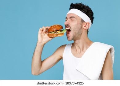 Young Sporty Fitness Man With Skinny Body Sportsman In Headband Shirt Towel Hold Eating American Classic Fast Food Burger Isolated On Blue Background Studio. Workout Gym Sport Motivation Concept