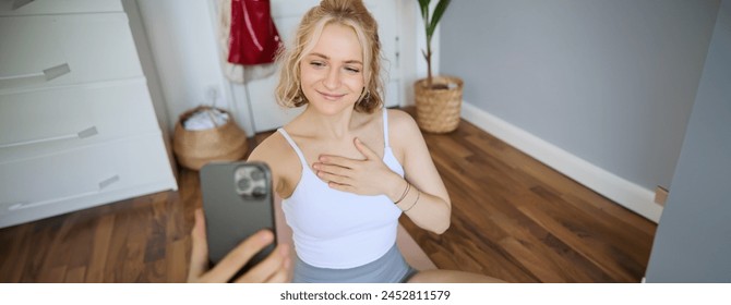 Young sporty fitness trainer, yoga instructor does live stream, takes selfies on smartphone while sitting on rubber mat at home, recording her workout training session. - Powered by Shutterstock