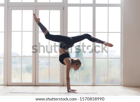 Young sporty flexible woman practicing yoga, doing handstand pose with splitted legs, indoor full length, yoga studio, free space