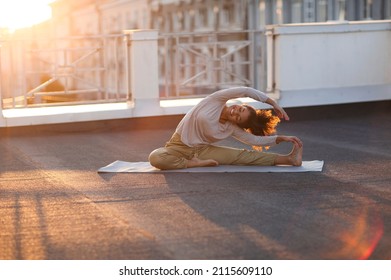 Young sporty happy african-american woman in sportswear doing stretching exercises while sitting on yoga mat on house roof in early morning, joyful fit female exercising at sunrise - Powered by Shutterstock