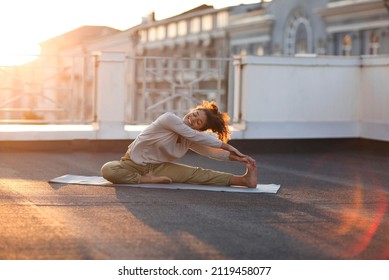 Young sporty happy mixed race woman in sportswear doing stretching exercises while sitting on yoga mat on house roof in early morning, joyful fit female exercising at sunrise - Powered by Shutterstock