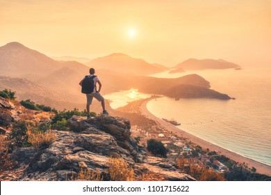 Young sporty man with backpack standing on the top of rock and looking at the seashore and mountains at sunset in summer. Scene with man, sea, mountain ridges and orange sky with sun. Oludeniz, Turkey - Powered by Shutterstock