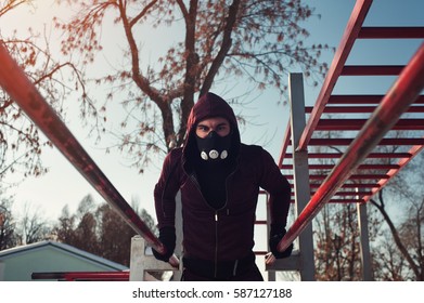 Young Sporty Man In High Altitude Mask Working Outdoors In Winter Background. Male Adult Exercising On Parallel Bars.