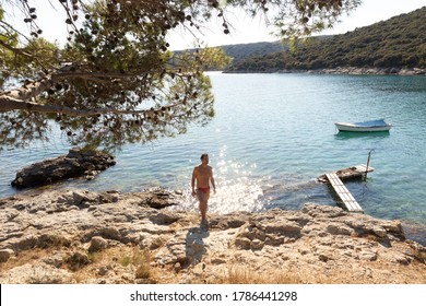 Young sporty man wearing red speedos tanning and realaxing on wild cove of Adriatic sea on a beach in shade of pine tree.