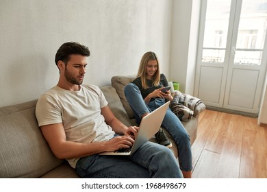 Young Sporty Man Works Hardly While His Girlfriend Watches Funny Content, Couple Sits On The Sofa With Plaid, Isolated Over The White Wall, Side View , Sitting In Front Of Balcony