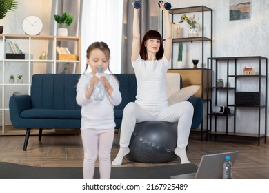 Young Sporty Mom Sitting On A Fitness Ball And Doing Sports Exercises With Dumbbells With Her Little Charming Girl In Sportswear Drinking Water From A Bottle While Standing After A Morning Workout.