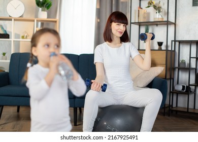 Young Sporty Mom Sitting On A Fitness Ball And Doing Sports Exercises With Dumbbells With Her Little Charming Girl In Sportswear Drinking Water From A Bottle While Standing After A Morning Workout.