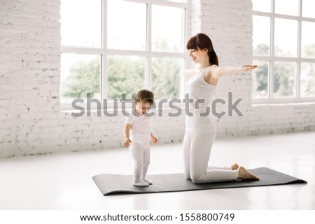 Young sporty mother and baby girl wearing white working out on the mat, exercising together, parent and child healthy development, game playing, fitness and relaxation. Healthy lifestyle concept