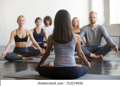 Young sporty people practicing yoga lesson with instructor, sitting in Sukhasana exercise, doing Easy Seat pose, working out, full length, studio, rear view at the female teacher. Wellbeing concept 