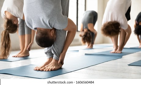 Young sporty people wearing sportswear practicing yoga at group lesson, standing in uttanasana pose on mats close up, doing forward bend, head to knees exercise, stretching in modern yoga studio