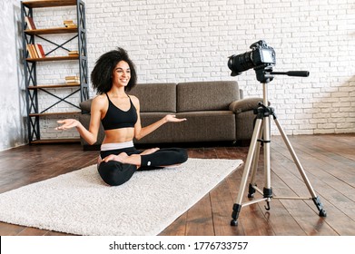 Young sporty slim mixed-race woman yoga coach records video online training yoga. An african-American girl sits in lotus pose and looks into camera in front of her and explains something - Powered by Shutterstock