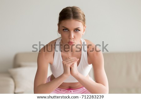 Young sporty slim woman doing squat morning exercise, practicing breathing correctly during working out home routine, looking at camera, holding hands together exhaling on muscular exertion, portrait
