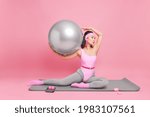 Young sporty slim woman dressed in activewear holds fitball poses on karemat excercises for healthy body spends free time at fitness studio isolated on pink background. Wellness aerobics workout