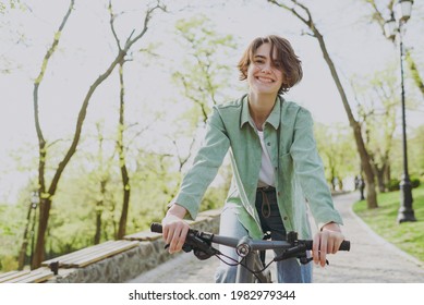 Young sporty smiling happy woman 20s wear casual green jacket jeans riding bicycle bike on sidewalk in city spring park outdoors, look camera. People active urban healthy lifestyle cycling concept. - Shutterstock ID 1982979344
