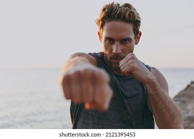 Young sporty strong athletic toned fit sportsman man in sports clothes warm up training boxing clench fist look camera at sunrise sun dawn over sea beach outdoor on pier seaside in summer day morning. - Powered by Shutterstock