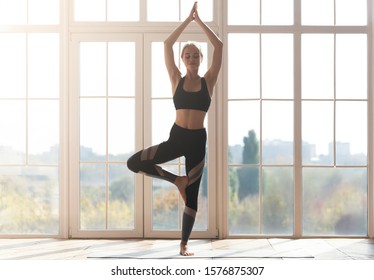 Young sporty woman doing yoga, practicing tree pose over big panoramic window, morning exercising, copy space