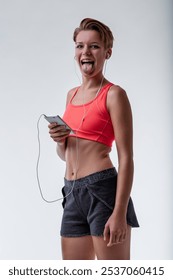 Young, sporty woman enjoys music with earphones, holding her phone and playfully sticking out her tongue on a white background. Her carefree expression exudes happiness and vitality