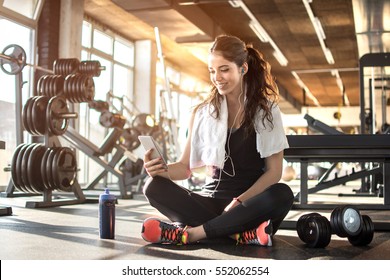 Young Sporty Woman Listening To Music On Smartphone In Gym. Break After Hard Workout.