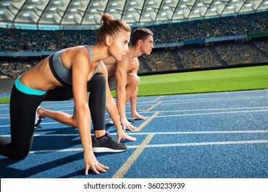 Young Sporty Woman And Man Are Ready To Run On Racetrack. Fit Well Formed People Are At Large Nice Modern Stadium
