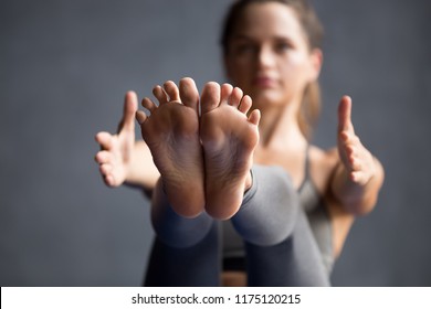 Young sporty woman practicing yoga, doing Paripurna Navasana exercise, boat pose, working out wearing sportswear grey pants, working out, indoor close up view of legs, yoga studio, focus on feet