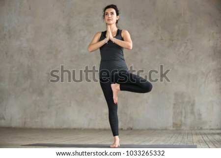 Young sporty woman practicing yoga, doing Vrksasana exercise, Tree pose, working out, wearing sportswear, black pants and top, indoor full length, gray wall in yoga studio