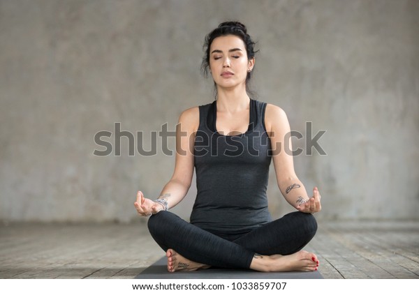 Young sporty woman practicing yoga, doing Sukhasana exercise, Easy Seat pose, working out, wearing sportswear, black pants and top, indoor full length, yoga studio