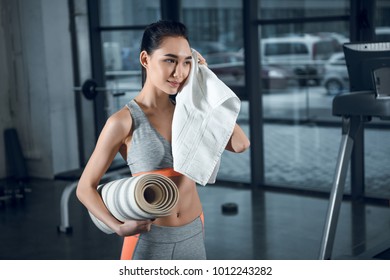 Young Sporty Woman With Rolled Yoga Mat Wiping Sweat With Towel After Exercises At Gym