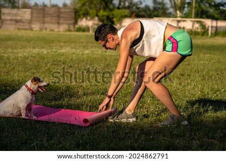 Young sporty woman with short black hair training outdoors on yoga mat on green grass in a park, activity with dog, jack russell terrier walk