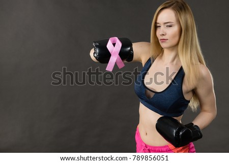 Young sporty woman wearing boxing gloves having pink ribbon tape, breast cancer symbol. Fighting with disease, feminine motivation. Studio shot on dark background.