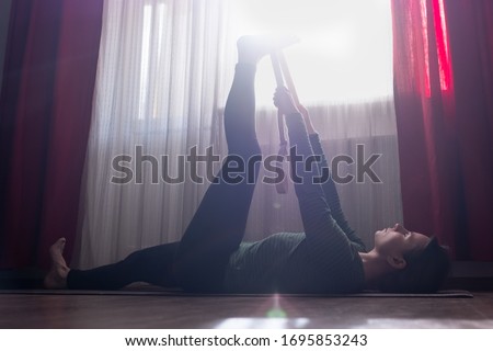 Young sporty woman working out at home , warming up using yogic belt, lying in yoga Supta Padangushthasana, One Leg Lift exercise, reclined variation of Extended Hand to Big Toe pose