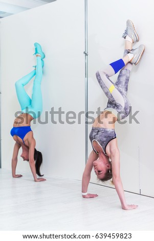 Young sporty women doing handstand exercise. Athletic girls standing in advanced downward-facing tree pose leaning against wall during yoga class indoors