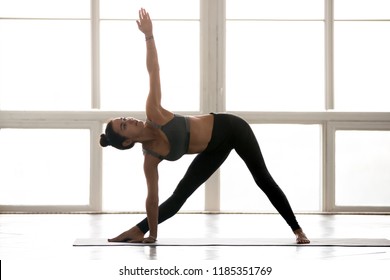 Young sporty yogi woman practicing yoga, doing Utthita Trikonasana exercise, extended triangle pose, working out, wearing sportswear, grey pants, top, indoor full length, at yoga studio