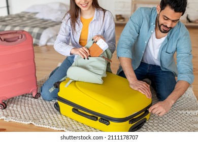 Young Spouses Packing Clothes In Suitcase And Woman Putting Passport With Tickets In Bag, Preparing For Vacation At Home, Free Space. Holidays Around The World Concept