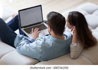 Young Spouses Sit On Sofa Relax Together At Home Using Computer, Buying Through E-commerce Retail E Services, Watch New Movie Online, Websurfing, White Mock Up Laptop Screen View Over Couple Rear View