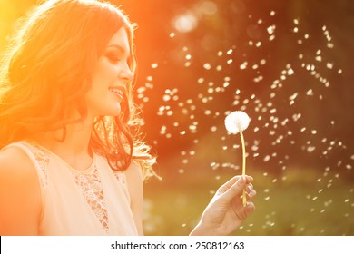 Young Spring Fashion Woman Blowing Dandelion In Spring Garden. Springtime. Trendy Girl At Sunset In Spring Landscape Background. Allergic To Pollen Of Flowers. Spring Allergy 