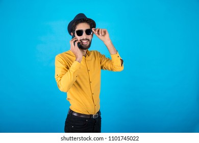 Young standing man talking on the phone touch the sunglasses on his face smiling on a blue background.