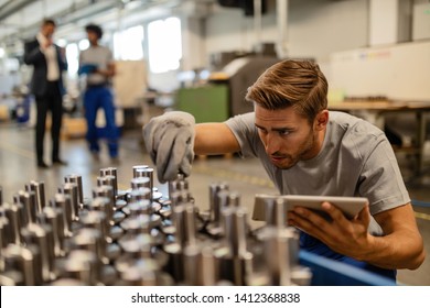 Young steel worker examining manufactured rod cylinder while using digital tablet in distribution warehouse. 