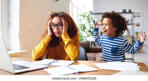 Young stressed afro american mother trying to work on laptop from home while little boy son playing around, making noise and disturbing her. Working mom and childcare concept