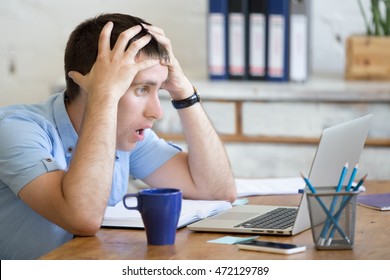 Young stressed businessman sitting with laptop and touching head with shocked facial expression. Surprised business man looking at laptop computer worried and amazed with open mouth and big eyes