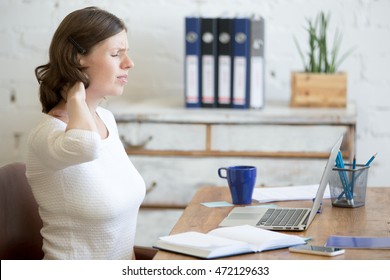 Молодая напряжённая деловая женщина сидит перед ноутбуком и держа шею с больным выражением. Деловая женщина чувствует боль, касаясь боли в шее, страдает от боли в шее после долгой работы на ПК