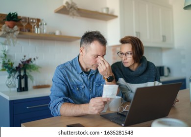 Young Stressed Caucasian Couple Facing Financials Troubles, Sitting At Kitchen Table With Bills, Checks And Laptop Computer Reading Document From Bank, Looking Frustrated And Unhappy