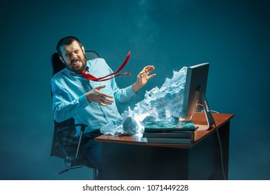 Young stressed handsome businessman working at desk in modern office shouting at laptop screen and being angry about e-mail spam. Collage with a mountain of crumpled paper. Business, internet concept