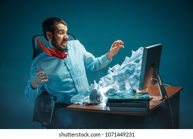 Young stressed handsome businessman working at desk in modern office shouting at laptop screen and being angry about e-mail spam. Collage with a mountain of crumpled paper. Business, internet concept