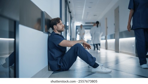 Young Stressed Male Doctor, Nurse or Surgeon in Scrubs Sitting Down Exhausted on a Hospital Hallway Floor, Trying to Rest, Fighting Stress and Fatigue in a Busy Medical Environment - Powered by Shutterstock