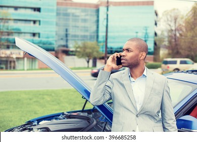 young stressed man having trouble with his broken car opening hood and calling for help on cell phone outside city urban background