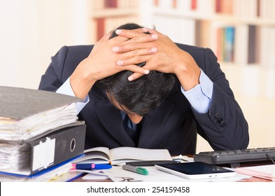 young stressed overwhelmed business man with piles of folders on his desk holding his head looking down