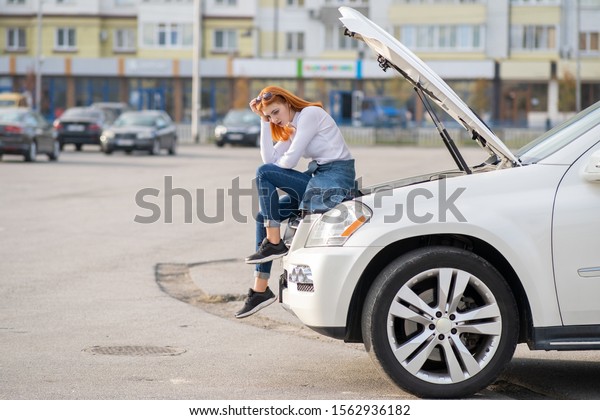 Young stressed woman driver near broken car\
with popped hood waiting for\
assistance.