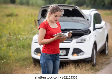 Young Stressed Woman Standing At Broken Car And Reading Owner Manual