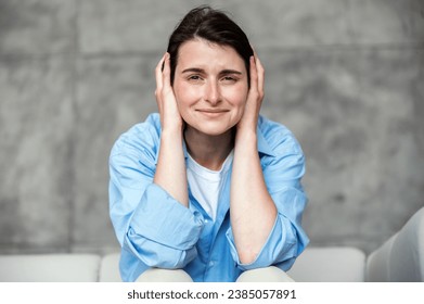 Young stressed woman wearing casual shirt, sitting on couch and closing ears with hands. Female don't want listening to unpleasant loud sounds or noise